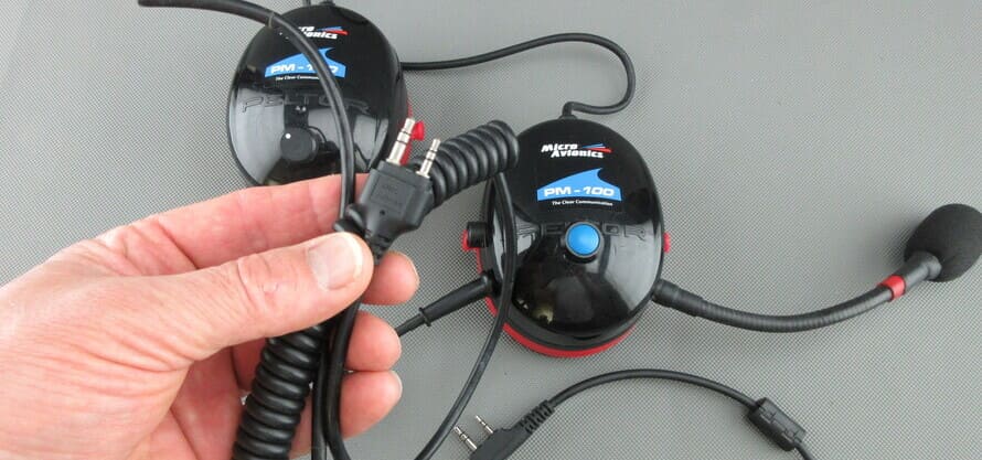 Ein MicroAvionics® Headset mit Funkgerätezuleitung, bei dem der Stecker für das Funkgerät in einer Hand gehalten wird.